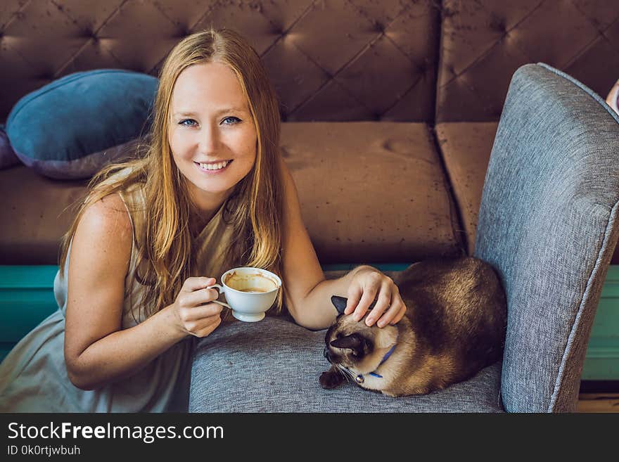 Young woman is drinking coffee and stroking the cat Against the backdrop of the sofa scratched by cats