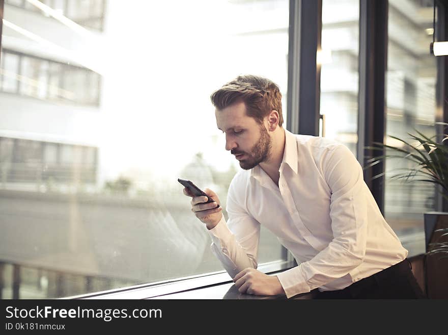 Photo of Man in White Dress Shirt Holding Phone Near Window