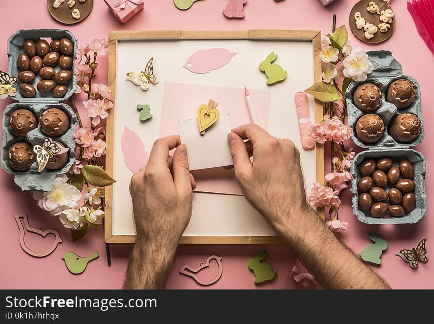 Easter composition, men`s hands hold a postcard, around are lined with various chocolate eggs, decorations and toys, top view flat