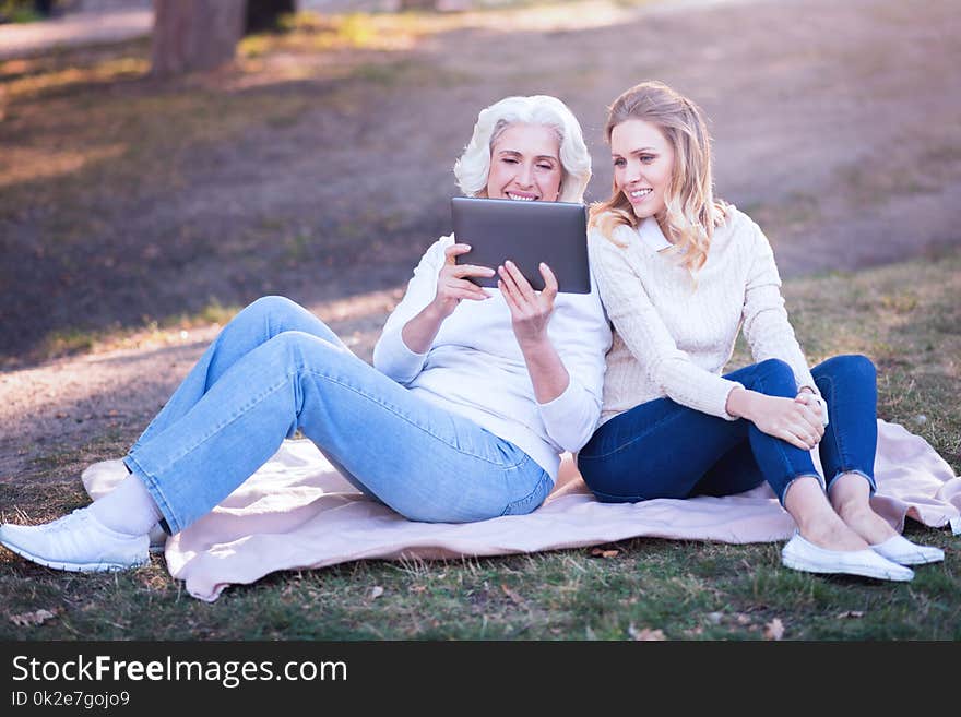 Spending family picnic. Stylish pleasant young women enjoying picnic and expressing happiness while using tablet with aged mother. Spending family picnic. Stylish pleasant young women enjoying picnic and expressing happiness while using tablet with aged mother