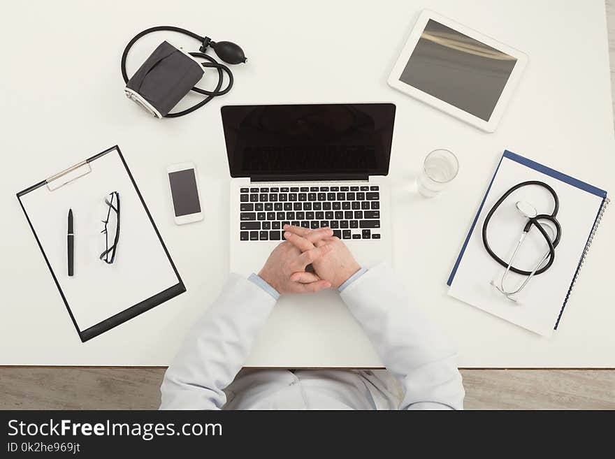 Doctor sitting at office desk with medical equipment and typing on his laptop with blank screen, top view, copy space. Doctor sitting at office desk with medical equipment and typing on his laptop with blank screen, top view, copy space