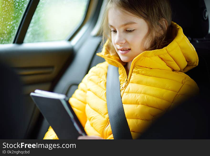 Adorable girl sitting in a car and reading her ebook on rainy autumn day. Child entertaining herserf on a road trip.