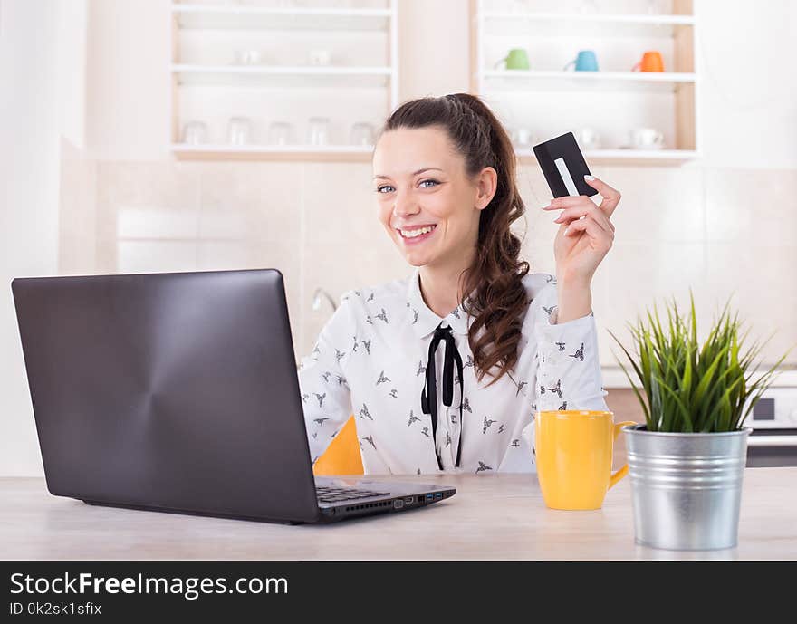 Happy young woman sitting at kitchen table with laptop and holding credit card. Online shopping and e-business concept. Happy young woman sitting at kitchen table with laptop and holding credit card. Online shopping and e-business concept