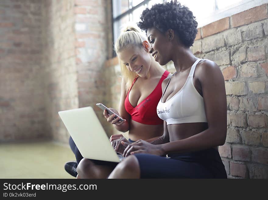 Two Women Wearing Red and White Sports Bras Sitting Near Brown Wall Bricks