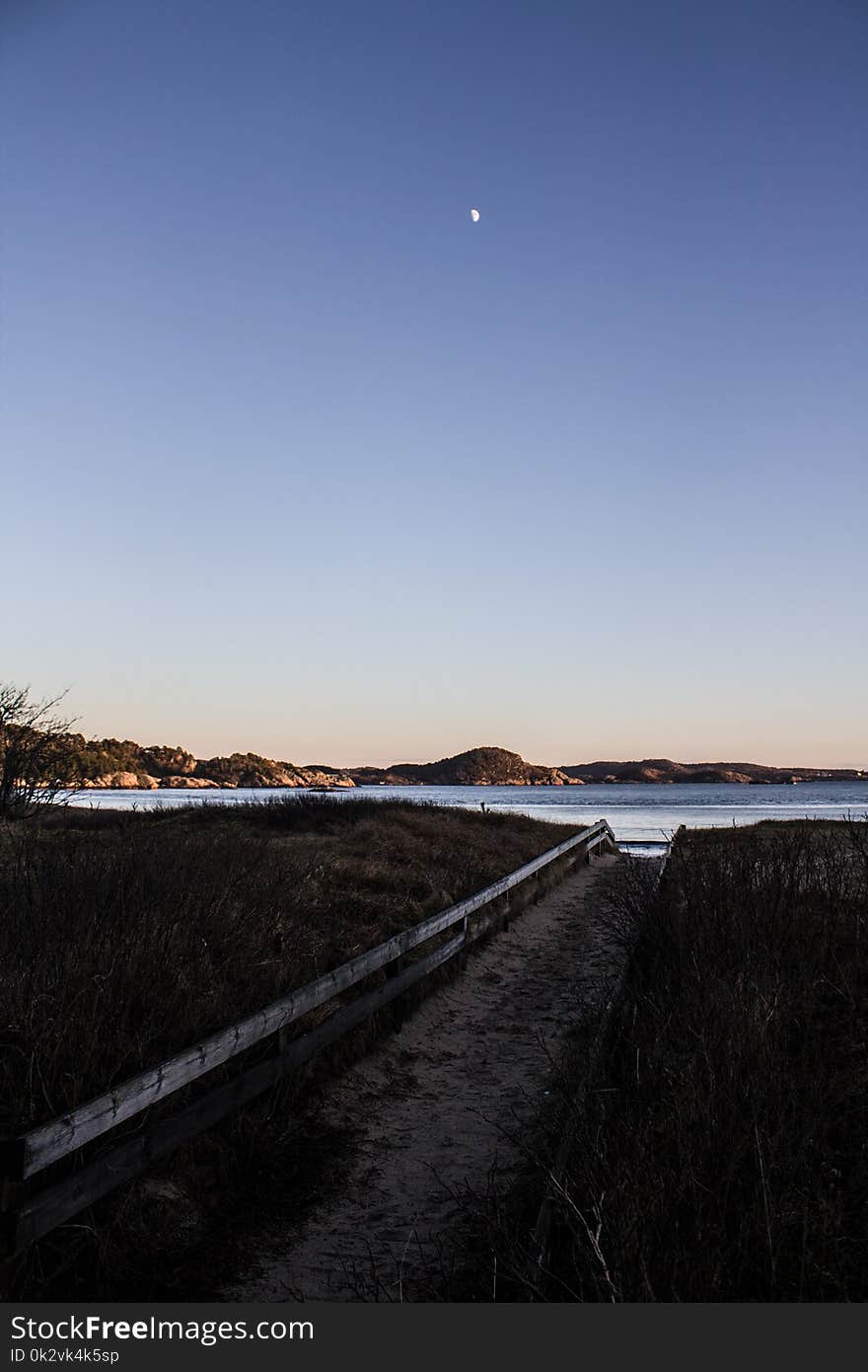 Pathway Lined With Green Grass Leading to Body of Water Under Blue Sky