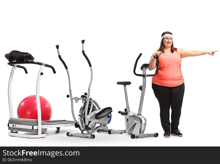 Full length portrait of an overweight woman standing by exercise machines and pointing isolated on white background