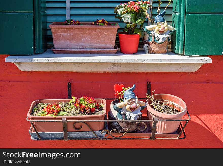 Daylight view to flowers in pot on balcony and windowsill. Little dwarf statues and sun shining on red painted building. Burano, Italy