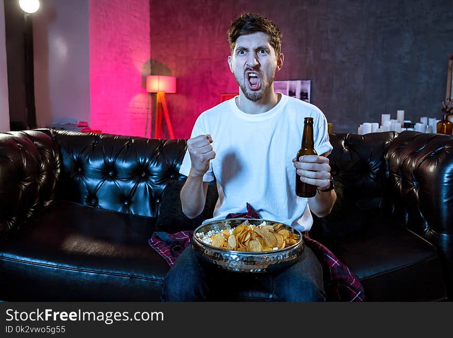 Young supporter man watching football game on television sitting at home couch with ball beer and chips in stress dejected and disappointed for failure shouting and complaining. Emotional fan watching the game of your favorite team