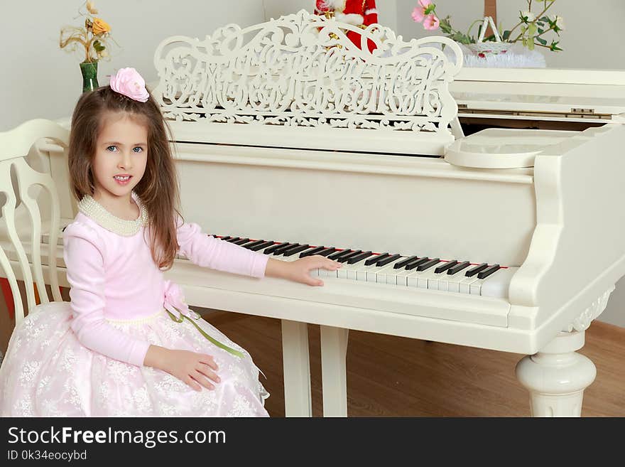 Cute little girl in beautiful pink dress and a rose in her hair.She put her hand on the keys of a vintage white piano. Cute little girl in beautiful pink dress and a rose in her hair.She put her hand on the keys of a vintage white piano.