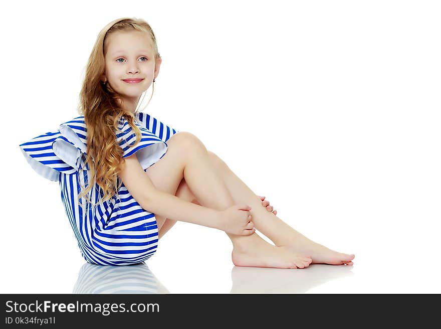 Adorable little blond girl in very short summer striped dress.She sits on the floor barefoot, turning sideways to the camera.Isolated on white background.