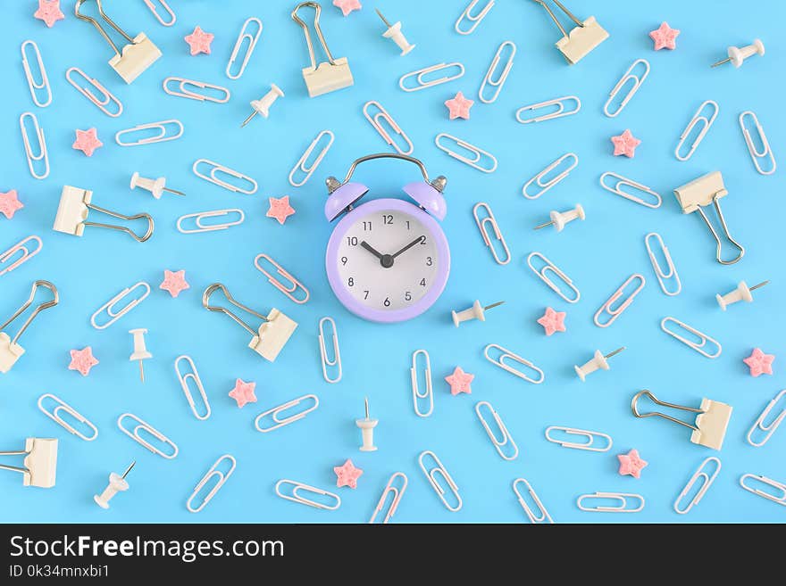 Stationery chaotically scattered on a blue background. White paper clips, clerical buttons and small pink stars in disorder . In the center is a small lilac alarm clock. Beautiful picture from the top.