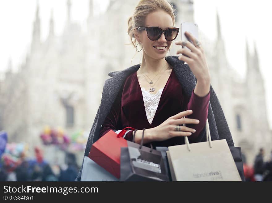 Woman in Maroon Long-sleeved Top Holding Smartphone With Shopping Bags at Daytime