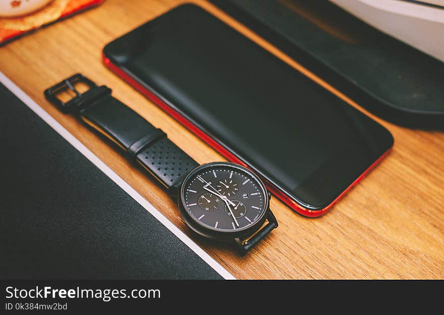 Red Smartphone Beside the Black Chronograph Watch on Brown Wooden Board