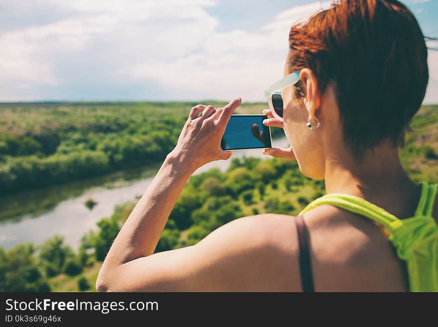 The girl is taking pictures on the phone. A woman takes a beautiful landscape on her cell phone. The brunette is traveling. A girl in glasses on the background of a river and a forest.