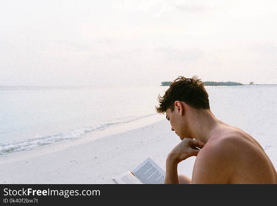 Topless Man Reading Book While Seating at Beach