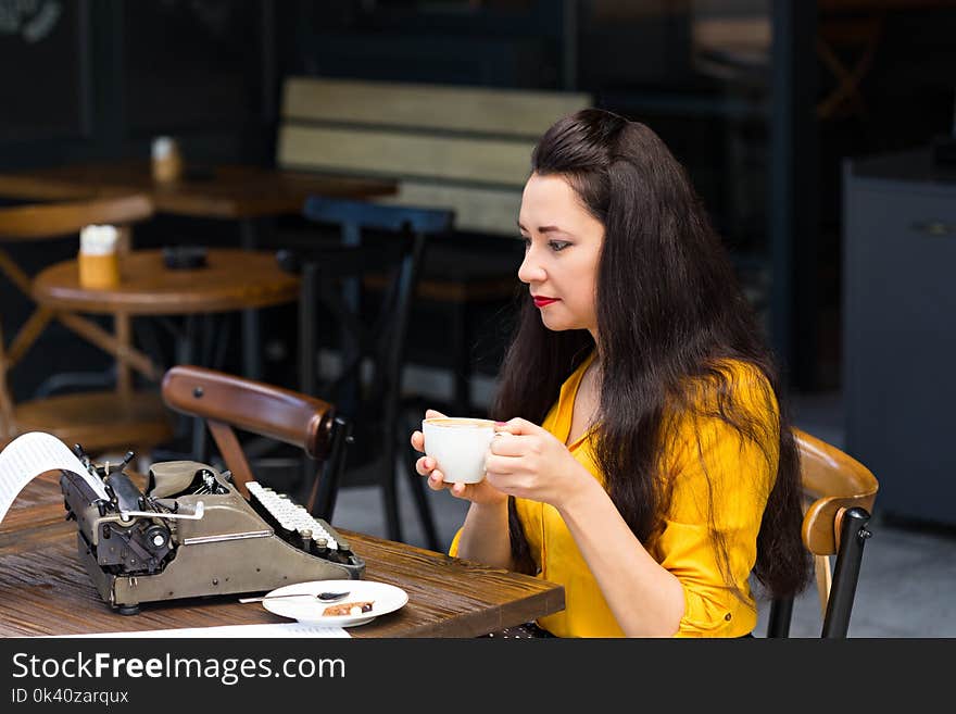 Freelance and writing concept. Writer female with dark long hair, wearing yellow shirt and working in a coffee shop using a retro typewriter. Freelance and writing concept. Writer female with dark long hair, wearing yellow shirt and working in a coffee shop using a retro typewriter.