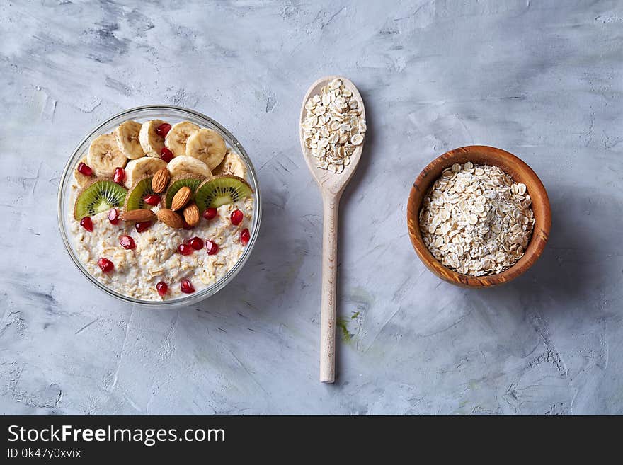 Diet breakfast oatmeal cereal with kiwi, banana, almond and pomergranate seeds and wooden bowl and spoon with oat flakes over white textured background, selective focus, close-up, top view. Healthy eating concept. Breakfast background. Diet breakfast oatmeal cereal with kiwi, banana, almond and pomergranate seeds and wooden bowl and spoon with oat flakes over white textured background, selective focus, close-up, top view. Healthy eating concept. Breakfast background.