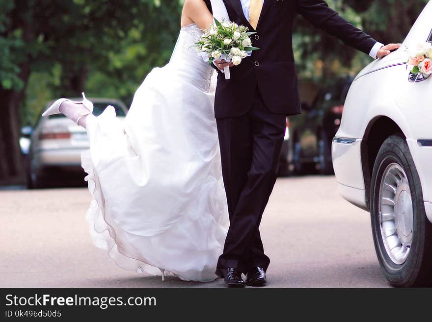 Bride and groom near white limousine, joyful, with wedding bouquet. Female leans on the male, playfully raising her leg