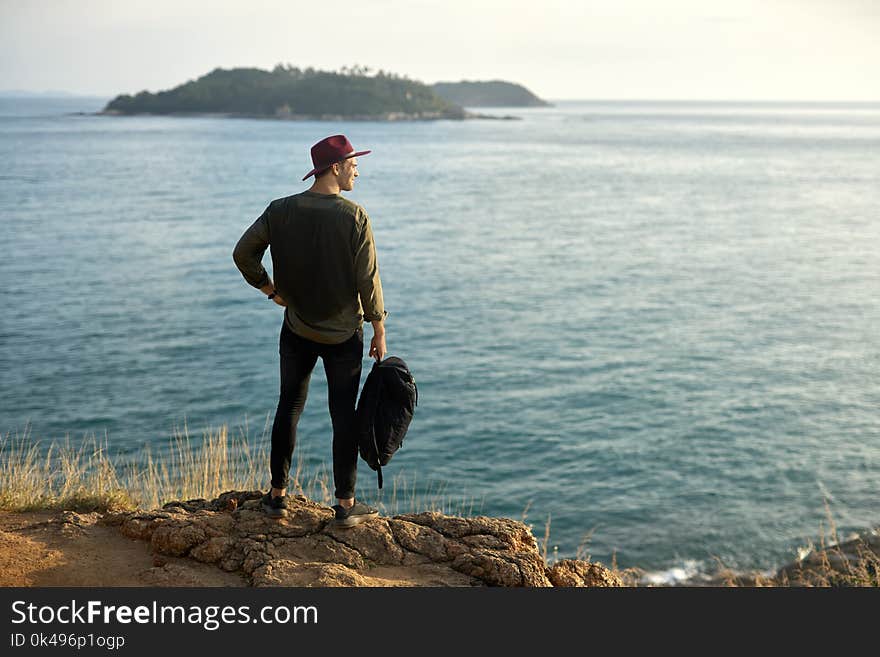 Handsome man with a black backpack stands on the cliff on the background of the green islands in the sea. He wears dark jeans and sneakers, olive shirt and crimson hat. Shoot from the back. Handsome man with a black backpack stands on the cliff on the background of the green islands in the sea. He wears dark jeans and sneakers, olive shirt and crimson hat. Shoot from the back.