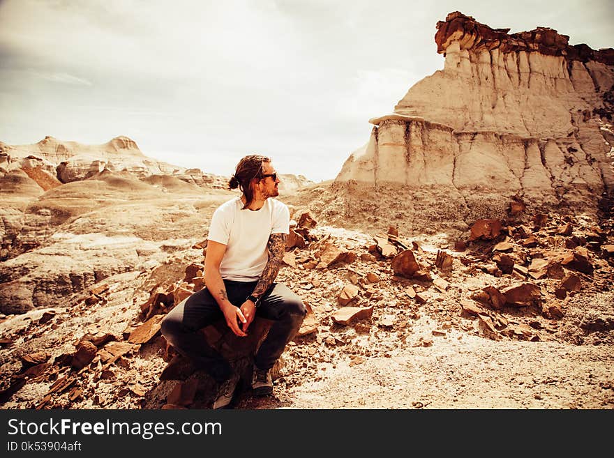 Man in White Crew-neck T-shirt and Black Pants Sitting on Boulder Near Cliff