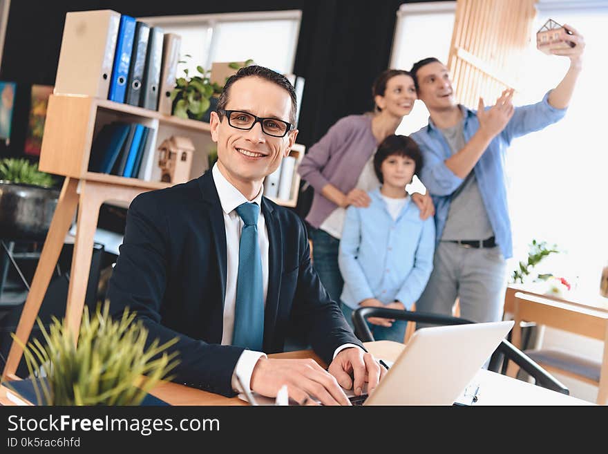 Realtor sitting at desk in office. Realtor is working on laptop with family in background.