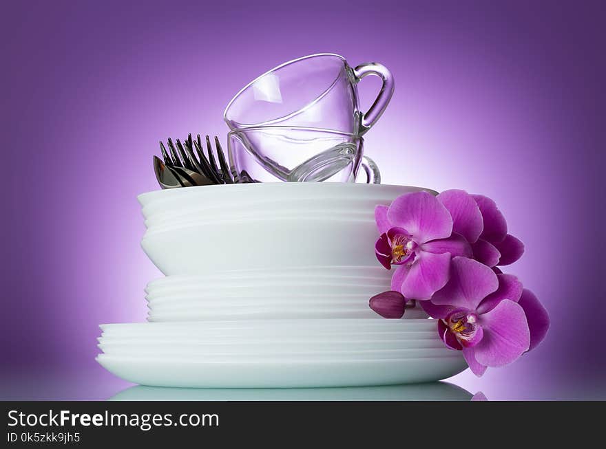 Stack of clean plates, cutlery and cups are top, near Orchid flower on lilac background