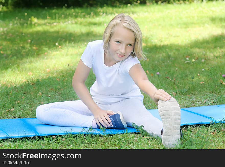 School-age girl in light clothes take sport exercise on a mat in the park. Outdoors workout.