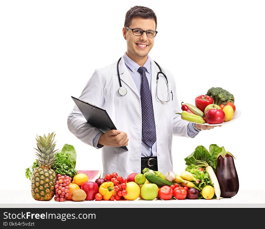 Doctor holding a clipboard and a plate behind a table with t and vegetables isolated on white background. Doctor holding a clipboard and a plate behind a table with t and vegetables isolated on white background