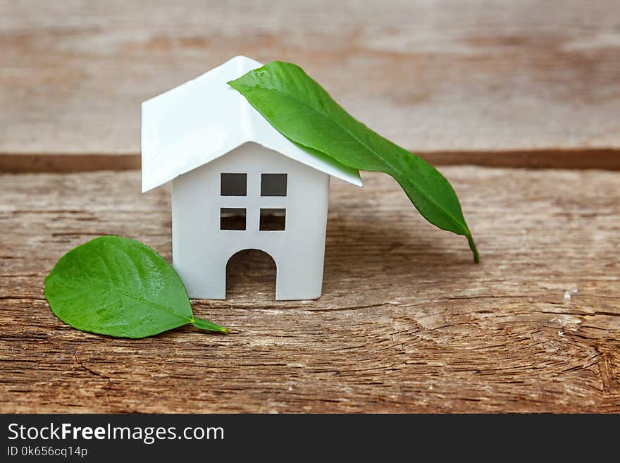 Miniature white toy model house with green leaves on wooden backgdrop. Eco Village, abstract environmental background. Real estate mortgage property insurance dream home ecology concept. Miniature white toy model house with green leaves on wooden backgdrop. Eco Village, abstract environmental background. Real estate mortgage property insurance dream home ecology concept