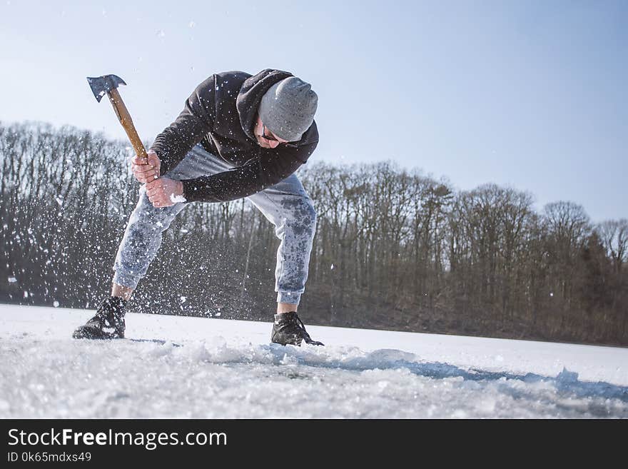 Man Wearing Black Hooded Jacket, Gray Knit Cap, Gray Pants, and Black Shoes Holding Brown Handled Axe While Bending on Snow