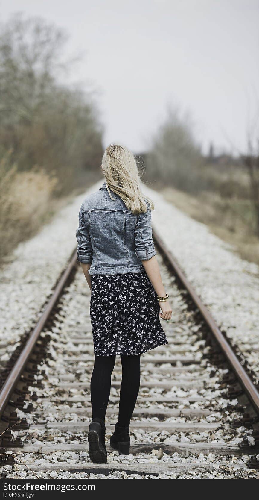 Woman in Blue Denim Jacket, Black Leggings and Black and White Dress Walking on Train Rail