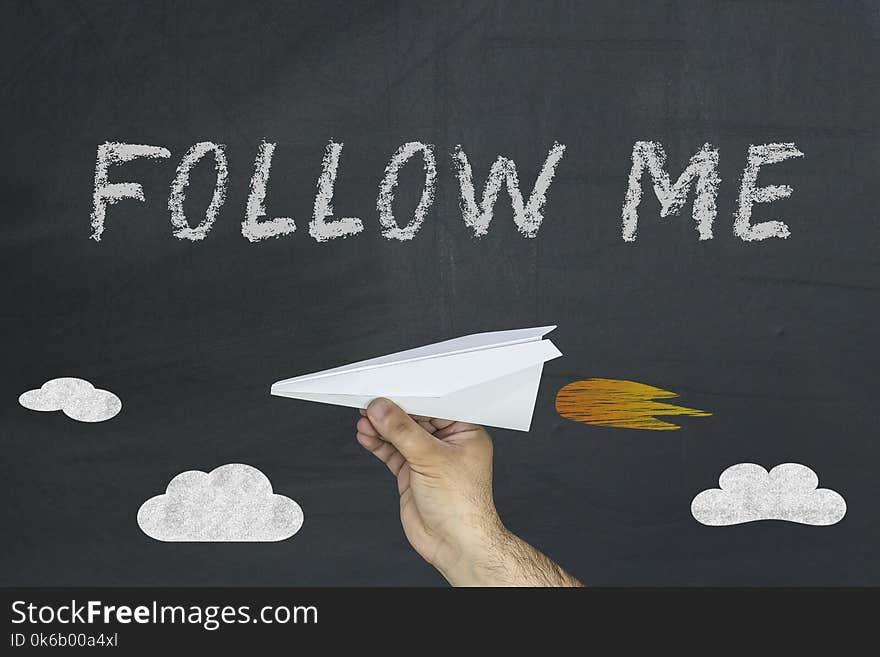 Human hand holding paper plane and Follow me words written on blackboard.