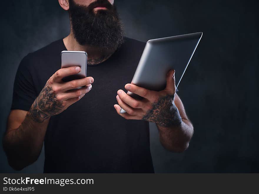 The tattooed, bearded hipster male holds tablet PC and smartphone on grey background. The tattooed, bearded hipster male holds tablet PC and smartphone on grey background.
