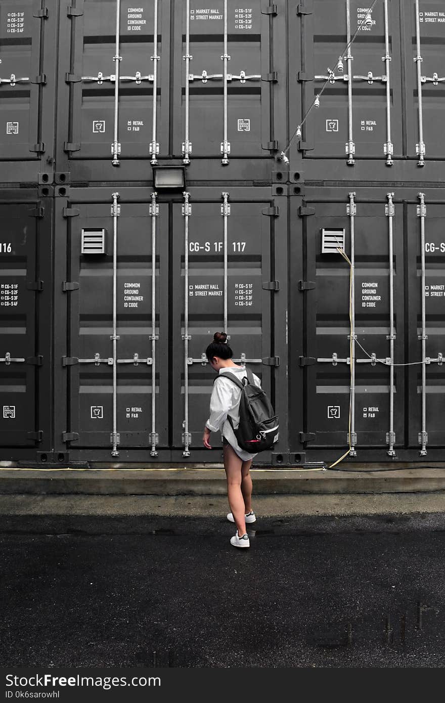 Photography of Woman in Gray Long Sleeve Shirt Carrying Black Backpack Standing in Front of Black Intermodal Container