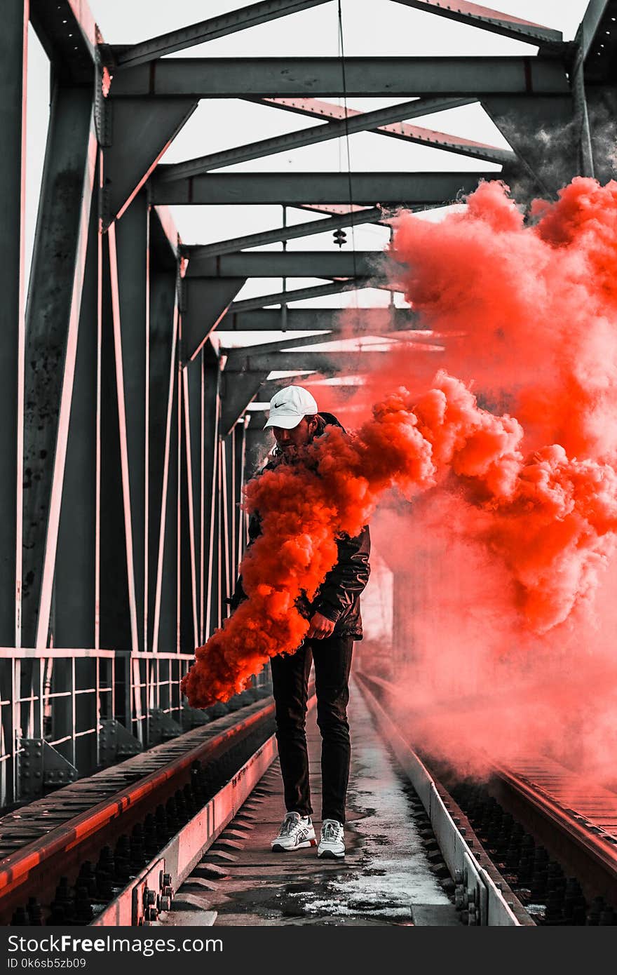 Man in Black Jacket Holding Red Smoke on Metal Bridge