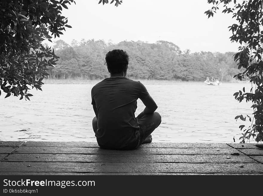 Gray Scale Photography of Man Sitting on Brown Wooden Floor Beside Body of Water