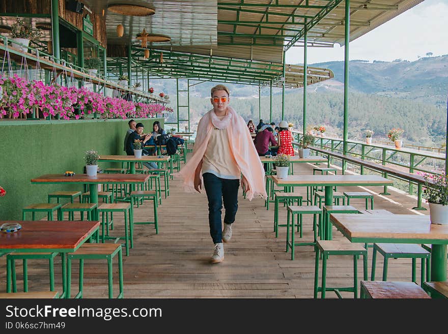 Man Wearing Yellow Undershirt, Green Denim Jeans, and Pair of White Sneakers Walking Between Tables and Chairs