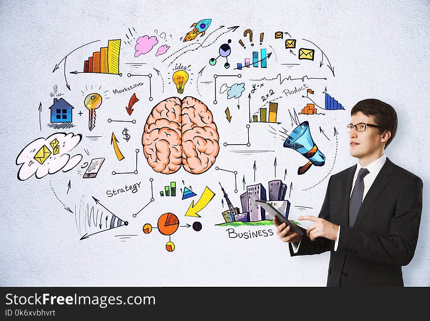 Handsome european businessman using tablet on concrete wall background with business brain sketch. Brainstorming, success and plan concept