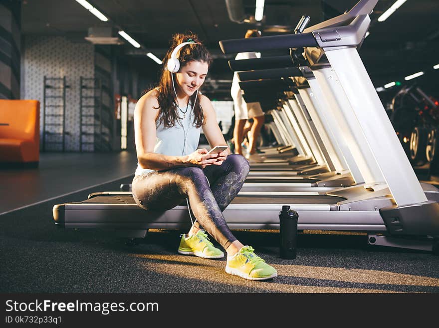 Beautiful young Caucasian girl sportswoman sitting, resting after training on treadmill against the backdrop of gym in sunny weath