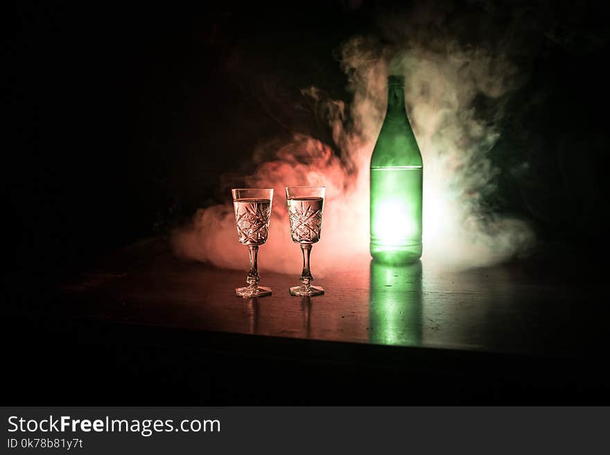 Two glasses of Vodka with bottle on dark foggy club style background with glowing lights (Laser, Stobe) Multi colored. Club drinks theme decoration. Empty space. Selective focus