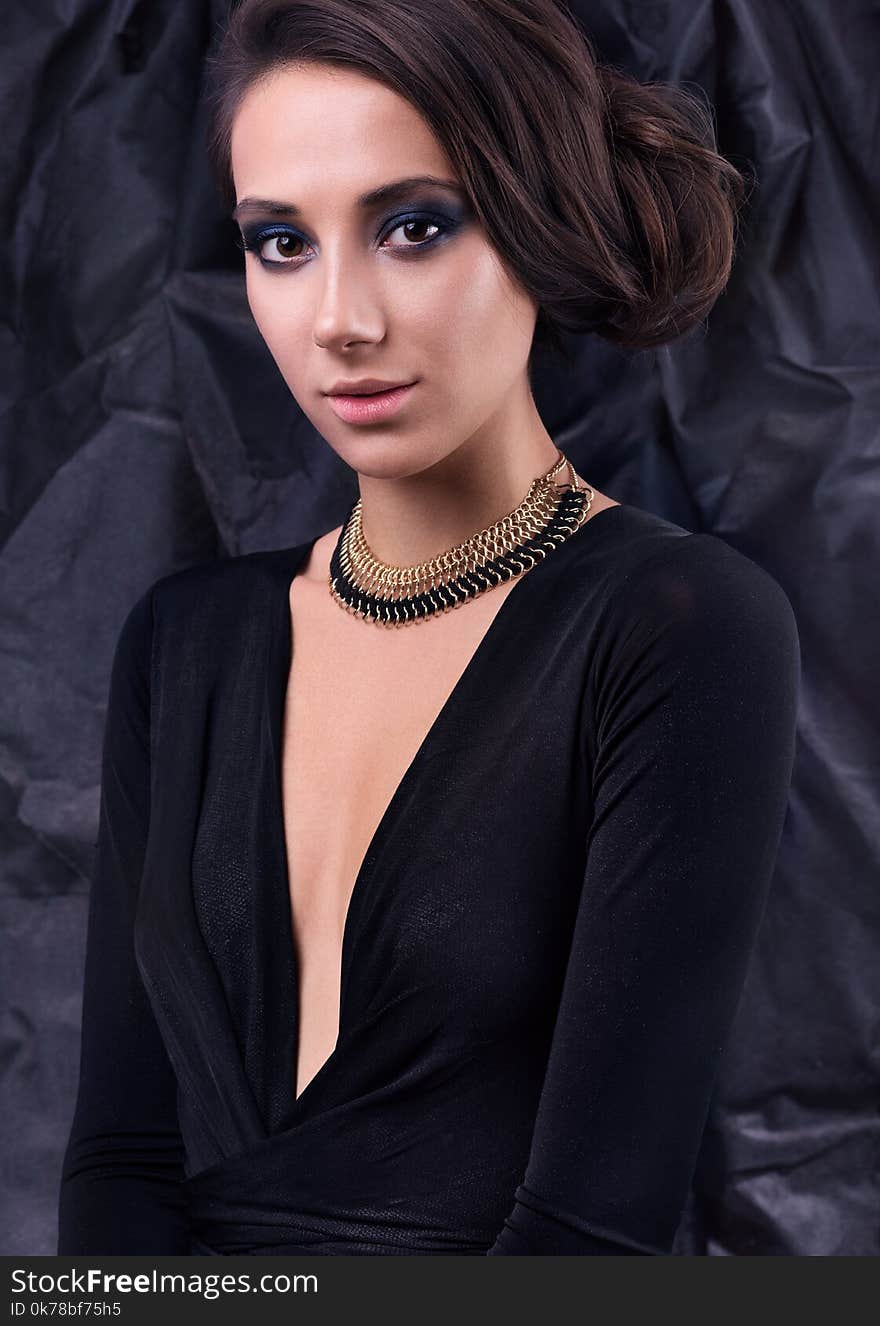 Studio portrait of young beautiful woman in evening dress. Necklace around her neck, hairstyle and makeup. Black dress and dark background