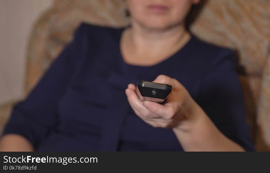 Close-up Of A Female Hand With A Remote Control On A Background Of A Woman In A Blue Dress