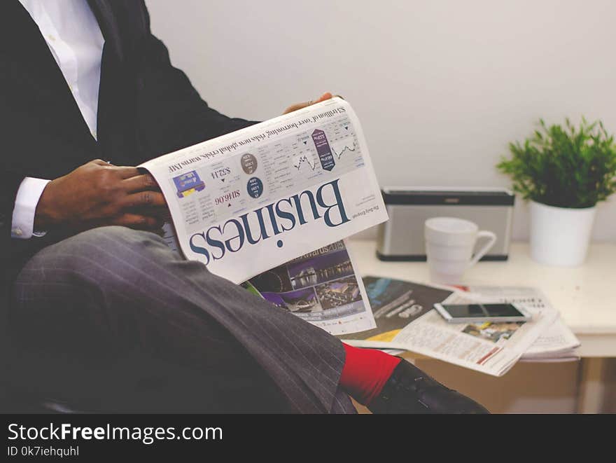 Man Reading Newspaper While Sitting Near Table With Smartphone and Cup