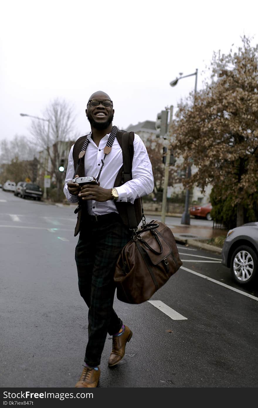 Man in White Button-up Dress Shirt Holding Camera While Smiling and Crossing Street