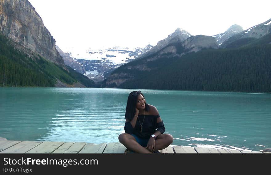 Woman With Black Off-shoulder Top Sitting on Wooden Dock Beside Blue Ocean With Mountain in Background