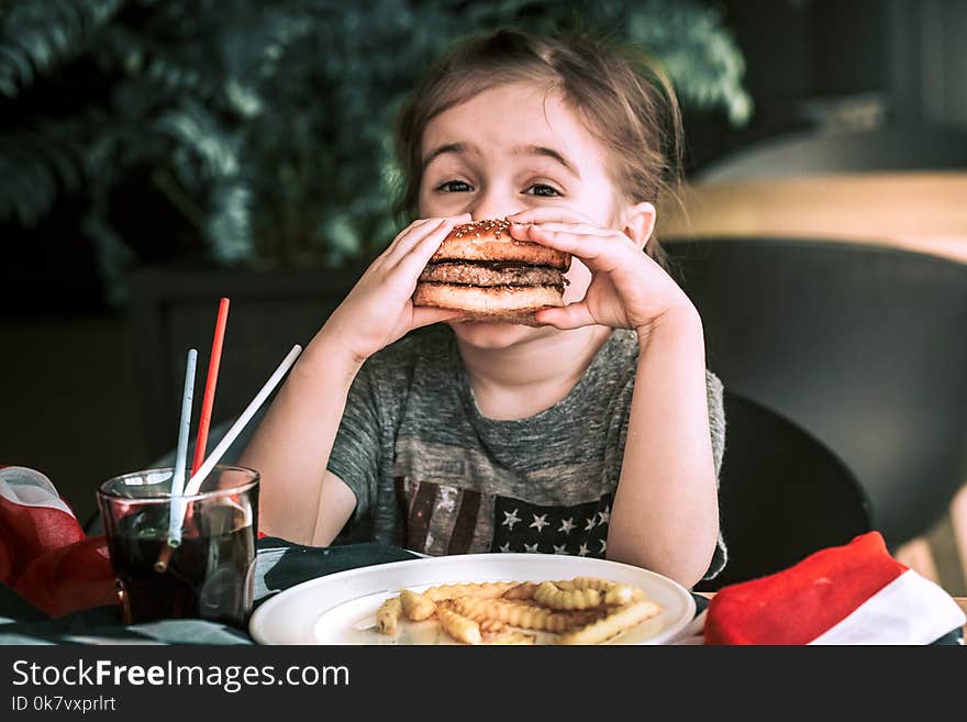 Little girl in T-shirt with American flag in cafe eating a burger and drinking a drink, concept of America Day and American food. Little girl in T-shirt with American flag in cafe eating a burger and drinking a drink, concept of America Day and American food