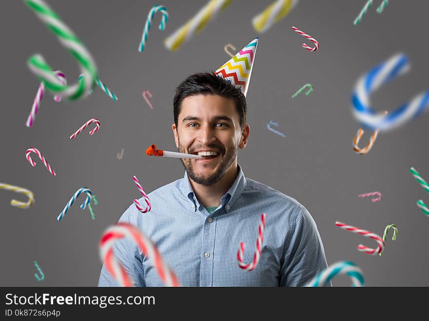 Handsome smiling young brunet guy with party cap making noise by red horn and standing under falling colorful candies. New year or birthday celebration. Handsome smiling young brunet guy with party cap making noise by red horn and standing under falling colorful candies. New year or birthday celebration