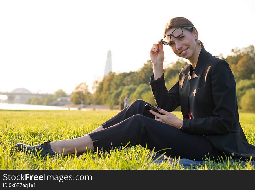 Young businesswoman in glasses using her smartphone and sitting on green grass at sunny day. Working outdoor, staying connected. Young businesswoman in glasses using her smartphone and sitting on green grass at sunny day. Working outdoor, staying connected