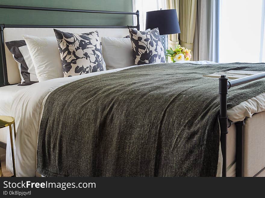 Black and white patterned pillows and black blanket on the bed with modern lamp and the beige curtain with warm light in shine through a blinds in bedroom. Black and white patterned pillows and black blanket on the bed with modern lamp and the beige curtain with warm light in shine through a blinds in bedroom.