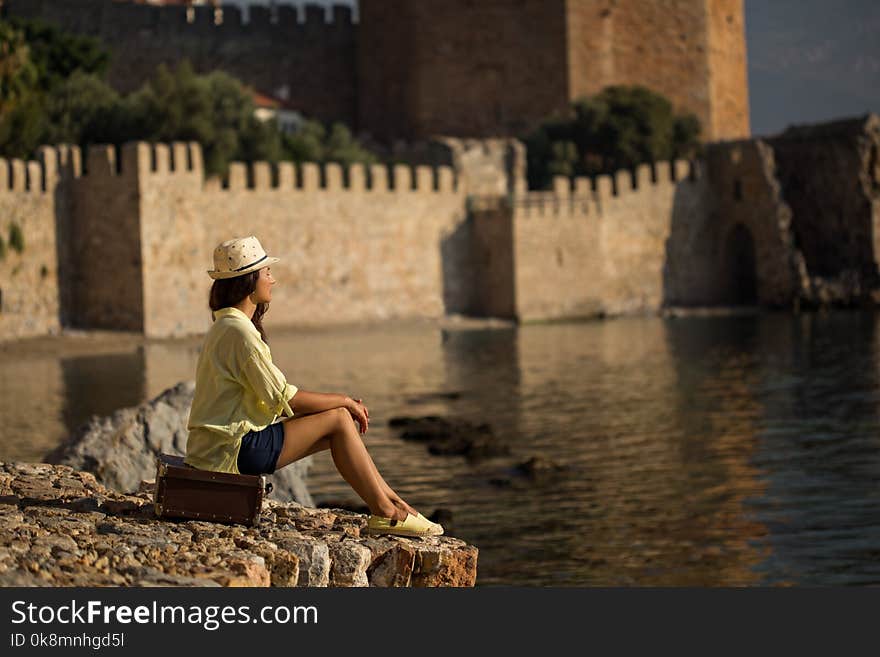 Sideview potrait of a solo traveller woman sitting on vintage suitcase by the sea and wearing dark blue shorts, yellow cotton shirt and hat. Copy space. Sideview potrait of a solo traveller woman sitting on vintage suitcase by the sea and wearing dark blue shorts, yellow cotton shirt and hat. Copy space.